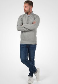 Hellgrauer Strickpullover mit einem Reißverschlusskragen und strukturiertem Rippenmuster, kombiniert mit blauen Jeans und weißen Sneakern. Lässige Passform und bequeme Gestaltung.