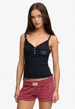 Jonge vrouw met lang bruin haar, gekleed in een zwart knoop-camisole en rood-wit gestreepte korte broek, staand tegen een effen achtergrond.