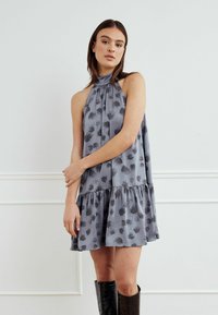 Siva haljina bez rukava s naboranim izrezom, višeslojnim rubom i tamnim cvjetnim uzorkom. Model nosi crne čizme do koljena. Glatka tekstura tkanine.