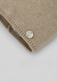 Tissu tricoté beige avec un motif texturé, comportant un bouton rond iridescent sur le bord du morceau plié.