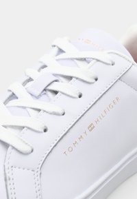 Biały skórzany sneaker z białymi sznurowadłami, z metalicznym logo "TOMMY HILFIGER" w kolorze różowego złota po boku i gładką teksturą.