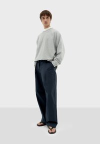 Sudadera gris de gran tamaño con mangas raglán, combinada con pantalones de pierna ancha de color azul oscuro. El modelo lleva chanclas negras.