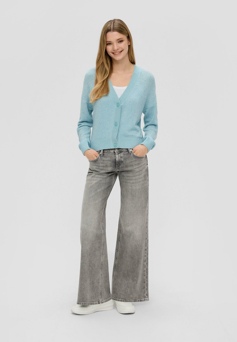 Jasnoniebieski sweter z dzianiny z dekoltem w serek, zapinany na guziki, z prążkowanymi mankietami, w połączeniu z szarymi spodniami o szerokich nogawkach i białymi trampkami.