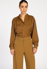 Vrouw in een bruine blouse met knopen en lange mouwen met manchet, en hooggewaaid brede broek met decoratieve sluitingen tegen een witte achtergrond.