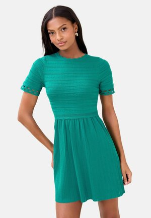 Kobieta w krótkim, zielonym, szmaragdowym, teksturowanym sukienka z dopasowanym górnym i luźną spódnicą, stojąca z jedną ręką na biodrze.