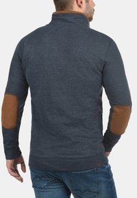 Mannen donkerblauwe trui met een hoge kraag, voorzien van bruine elleboogpatches, geribbelde manchetten en een gestructureerde stofafwerking.