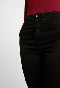 Close-up of a person wearing black high-waisted jeans and a red long-sleeve top against a plain light background.
