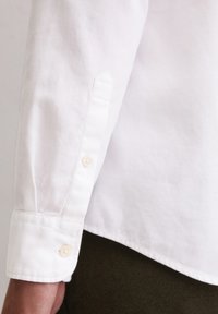 Witte katoenen shirt met een spitse manchett, voorzien van een enkele lichte knop bij de pols. Gladiatextuur en een strakke afwerking.