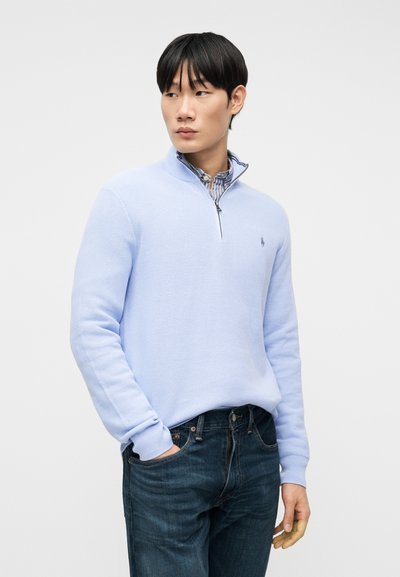 Νέος άνδρας φορώντας ένα ανοιχτό γαλάζιο πουλόβερ με φερμουάρ στο στήθος πάνω από ριγέ πουκάμισο, με σκούρο μπλε τζιν, στέκεται με το ένα χέρι στην τσέπη.