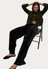 Zielony wełniany kardigan z złotymi guzikami, zestawiony z czarnymi aksamitnymi spodniami o szerokich nogawkach i czarnymi butami wsuwanymi, siedzący na czarnej składanej krześle.