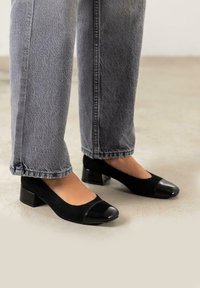 Zapatos de tacón negro con parte superior de ante y puntera de charol. Llevados con unos vaqueros grises, ligeramente cortos.