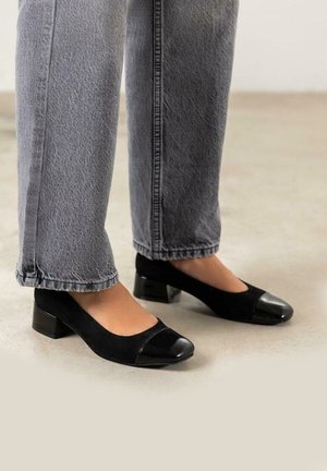 Zapatos de tacón negro con parte superior de ante y puntera de charol. Llevados con unos vaqueros grises, ligeramente cortos.