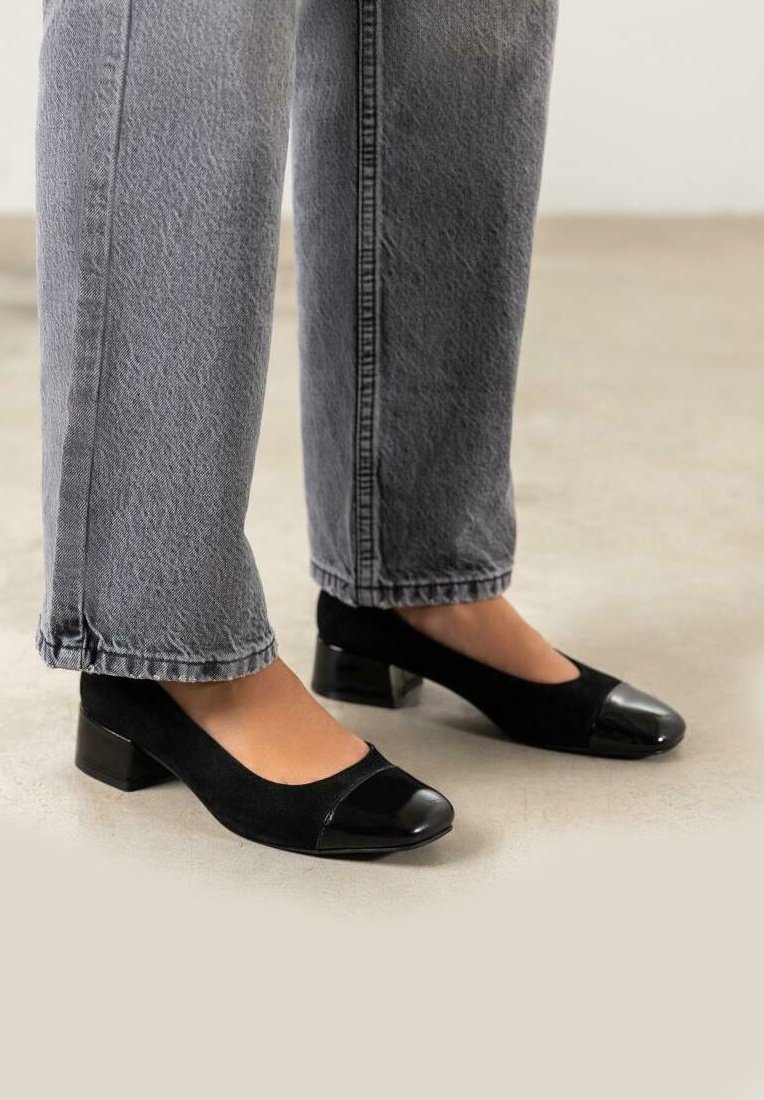 Zapatos de tacón negro con parte superior de ante y puntera de charol. Llevados con unos vaqueros grises, ligeramente cortos.