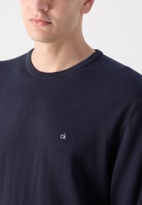 Tamsiai mėlynas apykaklės megztinis, pagamintas iš minkštos medžiagos, su mažuoju baltu „ck“ logotipu kairėje krūtinės pusėje. Lygi tekstūra ir kasdieniškas dizainas.