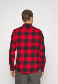 Chemise en flanelle à motif écossais rouge et noir avec un col boutonné, des manches longues et un ourlet droit, dotée de poignets avec fermetures à boutons.