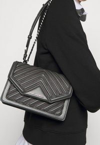 Borsa a tracolla in pelle trapuntata nera con motivo geometrico, dotata di chiusura a patta e tracolla a catena, esposta su una spalla vestita di nero.