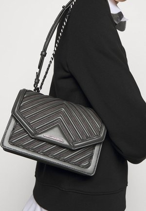 Schwarze, gesteppte Lederhandtasche mit geometrischem Muster, ausgestattet mit einem Klappenverschluss und einem Kettenriemen, der an einer schwarz gekleideten Schulter präsentiert wird.