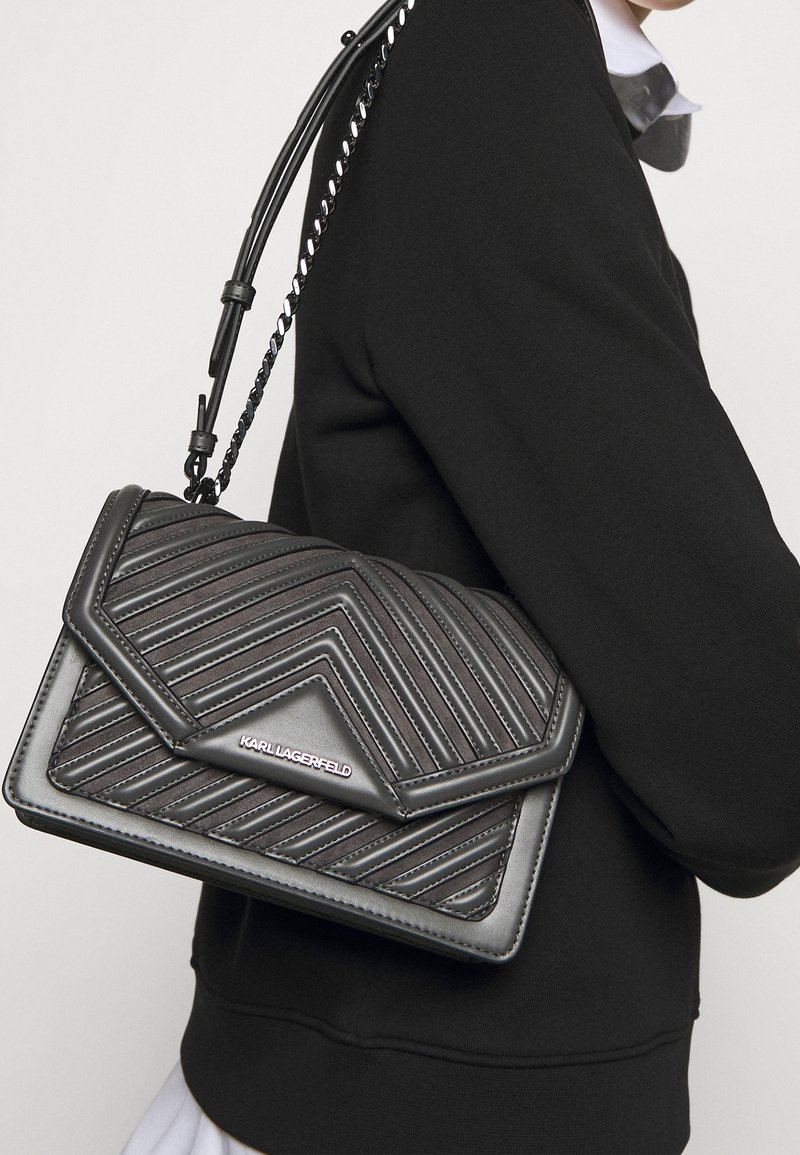 Borsa a tracolla in pelle trapuntata nera con motivo geometrico, dotata di chiusura a patta e tracolla a catena, esposta su una spalla vestita di nero.