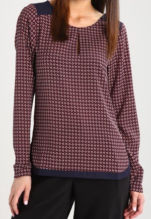 Femme portant une blouse à manches longues à motif bordeaux et blanc avec des accents bleu marine aux épaules et à l'ourlet, associée à un pantalon noir.