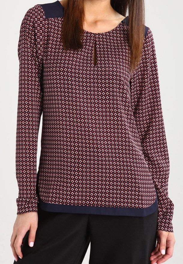 Femme portant une blouse à manches longues à motif bordeaux et blanc avec des accents bleu marine aux épaules et à l'ourlet, associée à un pantalon noir.