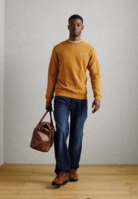 Felpa marrone, jeans blu e scarpe marroni. Tiene una borsa di pelle marrone. Design semplice e colori neutri su uno sfondo chiaro.