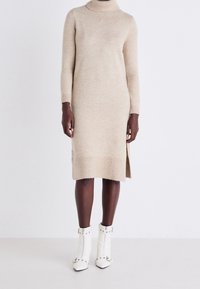 Personne à la peau foncée portant une robe en tricot beige à col roulé et à manches longues, avec des bottines blanches cloutées, se tenant devant un fond uni.