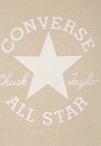 Béžová látka s bílým logem Converse All Star, které obsahuje velkou hvězdu a text "Chuck Taylor" po obou stranách.