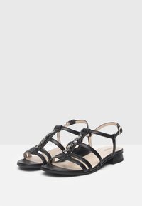 Sandali in pelle nera con più cinturini incrociati, dettaglio in catena metallica sulla parte anteriore, design con punta aperta e tacco basso.