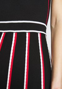 Robe en maille noire avec des rayures verticales blanches et rouges et une bande horizontale blanche à la taille, montrée sur le torse et le bras d'une personne.