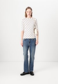 Chemise blanche avec des pois noirs et rouges, manches trois-quarts. Jean ajusté, légèrement évasé, en denim bleu clair. Chaussures noires.