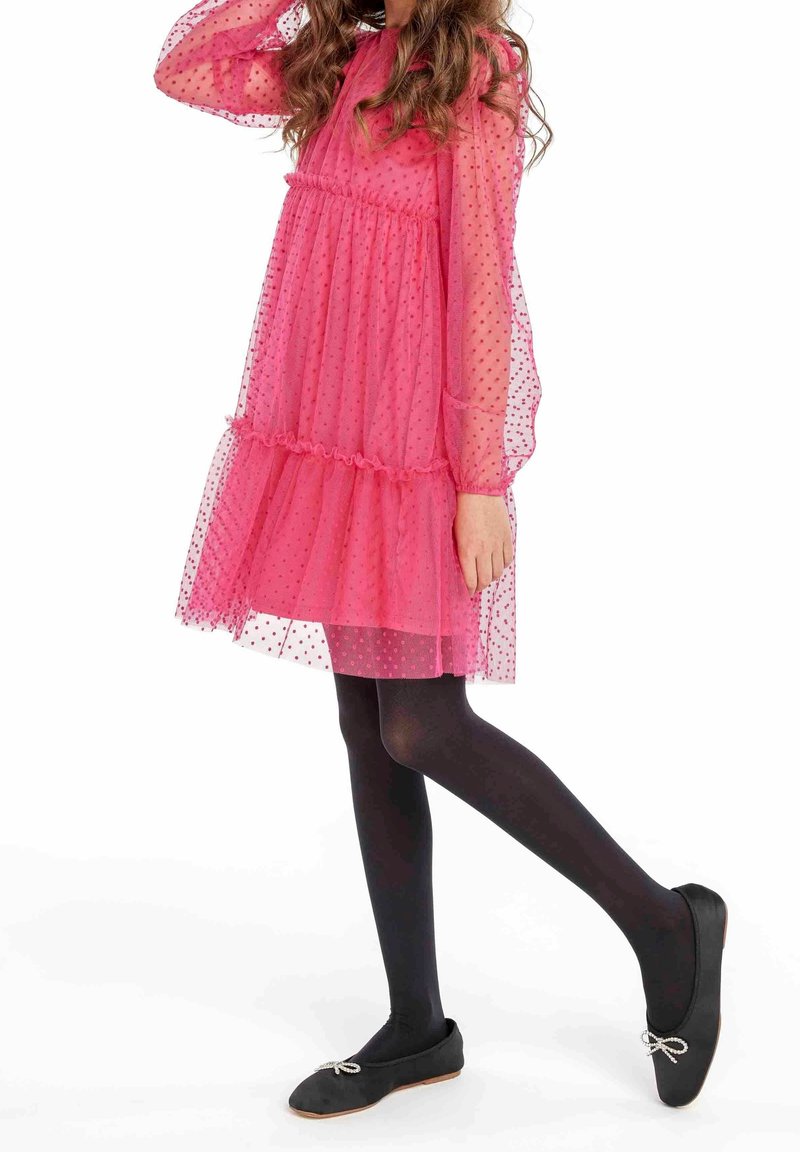 Fille portant une robe rose transparente à pois avec des manches longues, des collants noirs, et des ballerines noires avec de petits nœuds, posant avec une jambe levée.