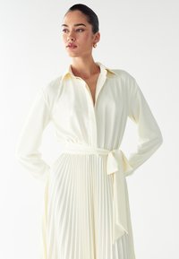 Langärmliges, knöpfbares Kleid in hellem Creme. Mit einem plissierten Rock, einem gebundenen Taillengürtel und einem klassischen Kragen. Geschmeidiger, fließender Stoff.