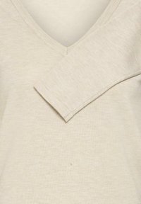 Haut beige à manches longues avec col en V, texture côtelée et couture oblique sur la manche gauche avec une finition soignée.