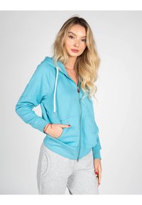 Felpa con zip in cotone di colore azzurro chiaro, dotata di tasca frontale, cordino regolabile e polsini a coste. Abbinata a pantaloni della tuta grigi.