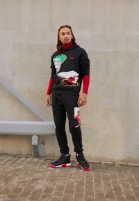 Black hoodie with graphic prints and red accents, paired with matching pants featuring patches. Black and red sneakers complete the outfit.