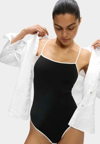 Bañador de una pieza negro con finas tiras blancas y un ribete blanco, usado debajo de una camisa blanca de botones abierta. Textura suave, diseño ajustado.