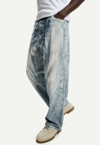 Jeans in denim azzurro chiaro con una vestibilità ampia, caratterizzati da zone sbiadite e dettagli decorativi in chiodi sulla parte inferiore delle gambe. Accentuati da tasche laterali.