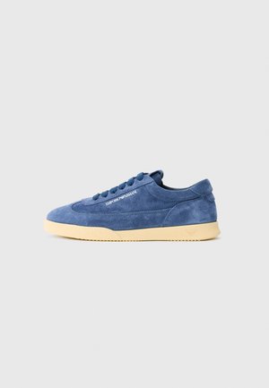 Blauwe suède lage sneakers met een crèmekleurige zool, veters en het 'EMPORIO ARMANI' logo op de zijkant, gefotografeerd op een effen witte achtergrond.
