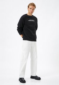 Zwarte sweatshirt met witte tekst, gecombineerd met witte cargo pants met zakken. Model draagt zwarte laarzen en staat op een effen achtergrond.