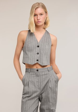 Frau mit blonden Haaren, die eine graue ärmellose Weste mit Nadelstreifen und passende hoch taillierte Hose trägt, mit den Händen in den Taschen vor einem einfarbigen Hintergrund.