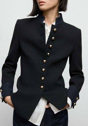 Blazer noir ajusté avec des boutons dorés, col haut et poignets contrastants. Présente une silhouette structurée et un tissu texturé.