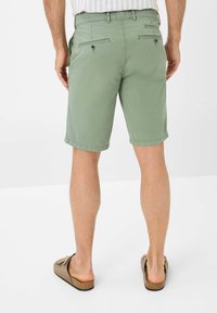 Pantaloni corti in cotone verde con taglio dritto, dotati di due tasche posteriori con bottoni e orli arrotolati. Indossati con sandali scivolati color cuoio.