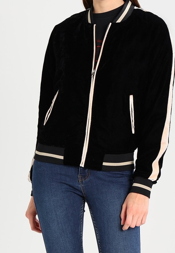 Giacca bomber nera con una texture morbida, caratterizzata da strisce crema sulle maniche e polsini e orlo a costine. Chiusura con zip e tasche incluse.