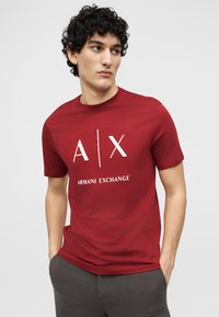 Νεαρός με σγουρά μαλλιά, φοράει κόκκινο μπλουζάκι Armani Exchange και σκούρο παντελόνι, κοιτάζοντας προς τα αριστερά με τα χέρια στις τσέπες.