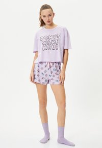 Crop top lavanda con grafica "DONUT WAKE ME UP", abbinato a pantaloni corti lavanda con un motivo a ciambella. Il modello indossa calzini lavanda abbinati.
