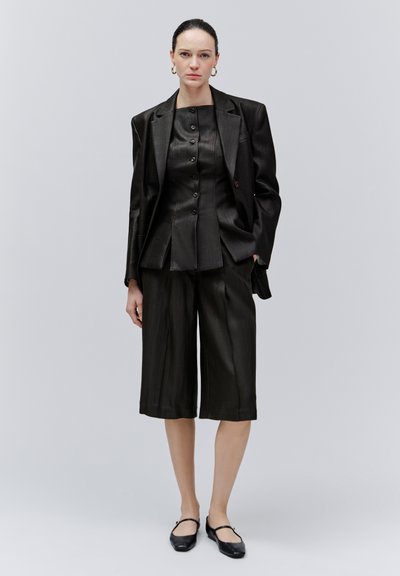 Femme debout portant un blazer noir oversize, un haut noir boutonné, un pantalon large noir style culotte, des ballerines noires et des boucles d'oreilles créoles dorées.