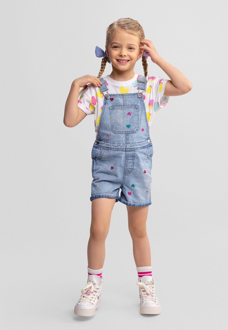 Fille souriante aux cheveux tressés portant une salopette en denim à motif de cœurs, un chemisier coloré à imprimé de cœurs, des chaussettes rayées et des baskets blanches.