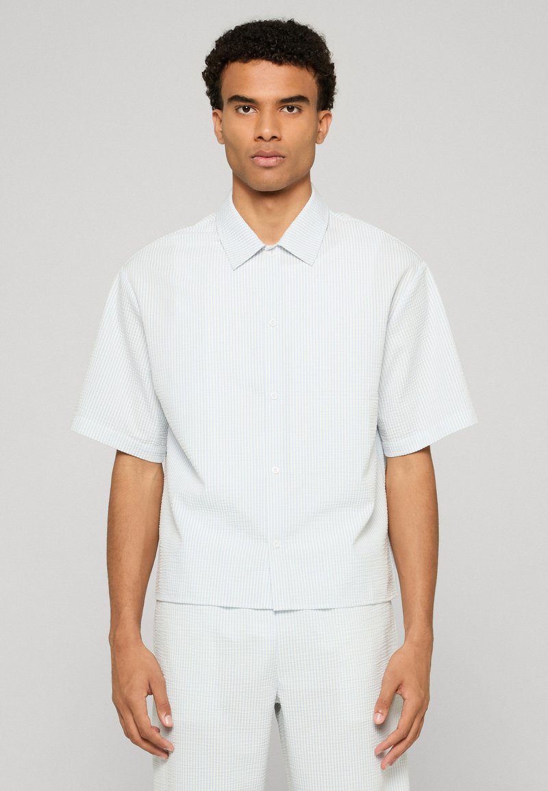 Jeune homme aux cheveux courts et bouclés portant une chemise à manches courtes boutonnée à rayures bleues claires et blanches ainsi qu'un pantalon assorti, debout immobile.