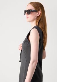 Black rectangular sunglasses with bold frames, worn by a model in a sleeveless dark dress, highlighted by sleek texture and subtle curves.