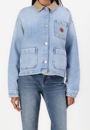 Personne portant une veste en jean bleu clair avec un col beige, trois poches avant, des boutons-pression et un écusson brodé sur la poche poitrine.